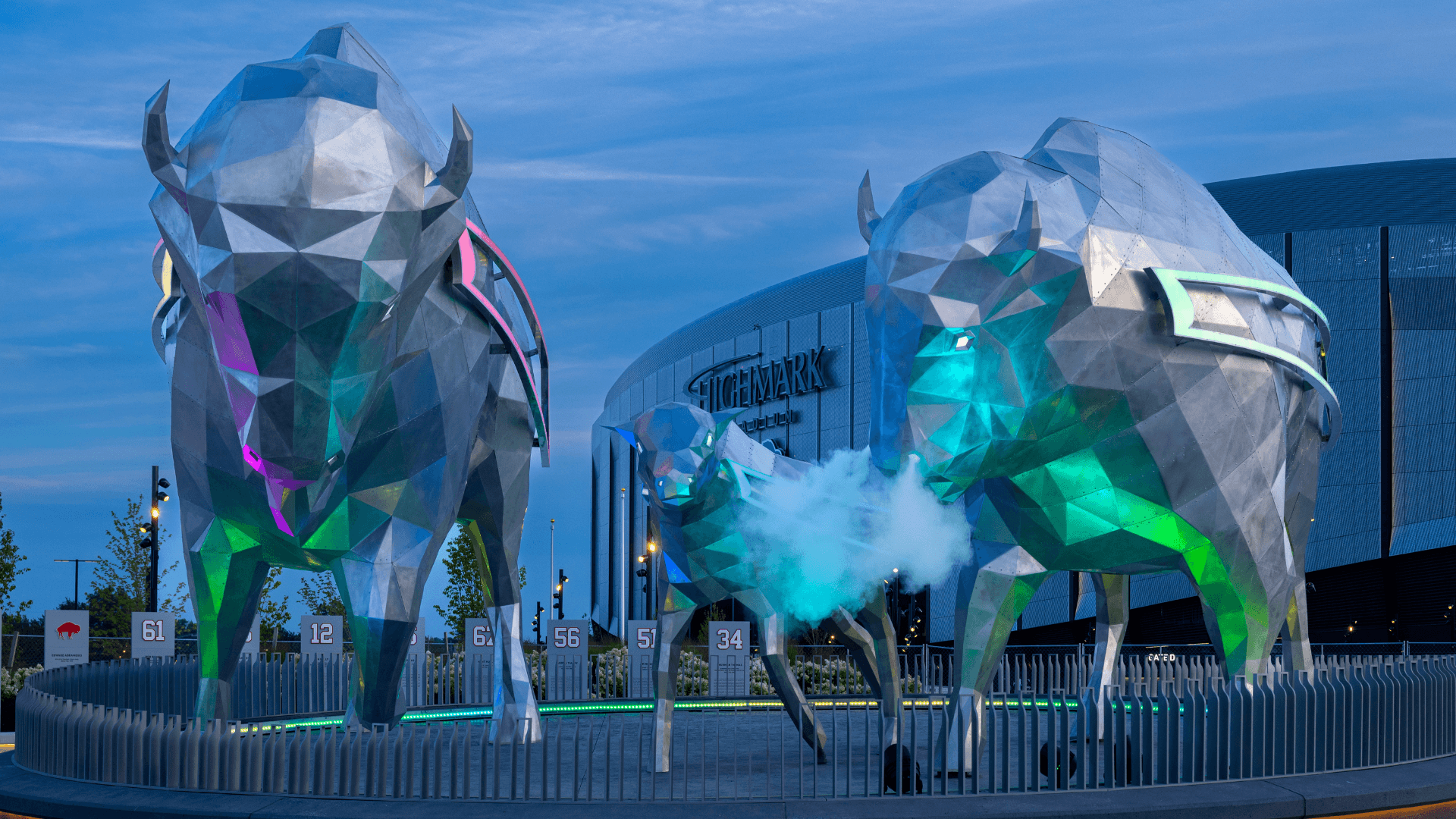 Three large, angular metallic bison sculptures with colorful lights stand outside a modern arena building during dusk. Mist rises from the smaller bison’s nostrils.